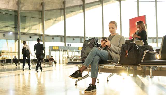 Woman with luggage sits at airport terminal looking at her phone.