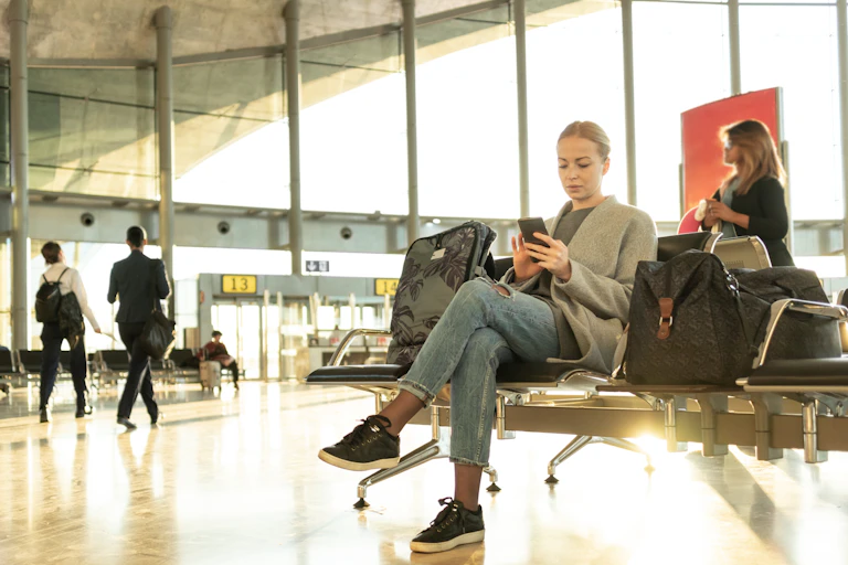 Woman with luggage sits at airport terminal looking at her phone.