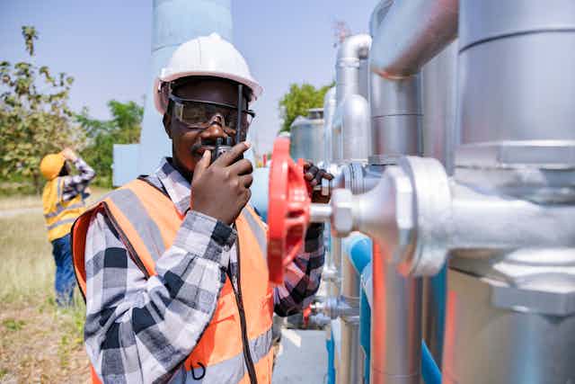 Man in hard hat and protective eyewear and reflective jacket speaks into a walkie-talkie while standing next to industrial equipment
