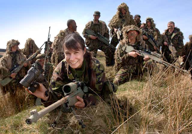 Soldiers at a sniper training course. A female solder holding a gun and binoculars, in the foreground and smiling at the camera. A number of male soldiers are in the background. They are outdoors, and all wearing military fatigues.