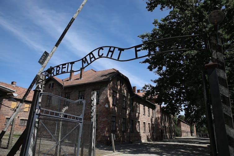 Modern photo of metal gate with words 'Arbeit macht frei' above it, leading to warehouse-like buildings.