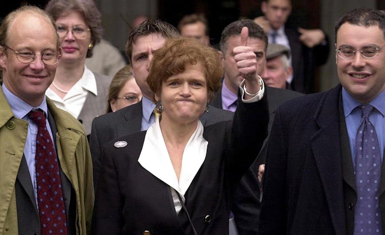 Smartly dressed woman gives thumbs up, flanked by two smiling men.
