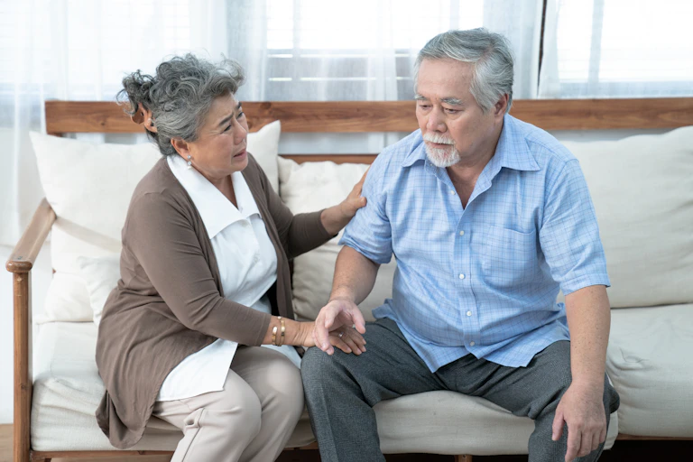 An older couple on a sofa; the woman looks concerned and is trying to assist the man