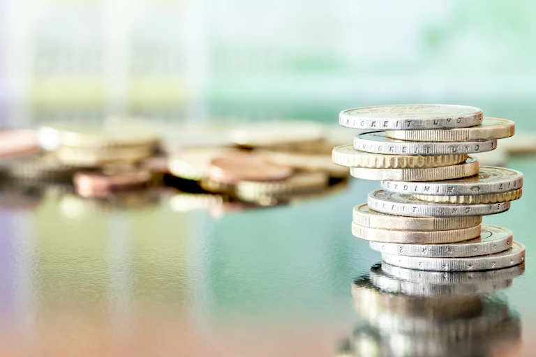 Coins are unevenly stacked in the foreground, with a pile of coins are sitting on a table in the background.