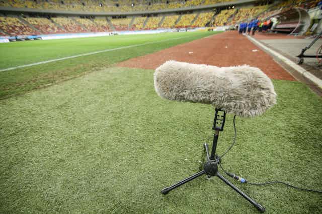 A microphone with a wind shield on a stand is placed on the sidelines of a grass playing field in a stadium