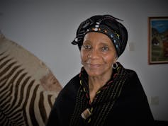 Uma mulher mais velha sorri levemente ao olhar para a câmera, olhos calorosos e vestidos de preto, um traje tradicional Xhosa com contas e uma pele de zebra na parede atrás dela.