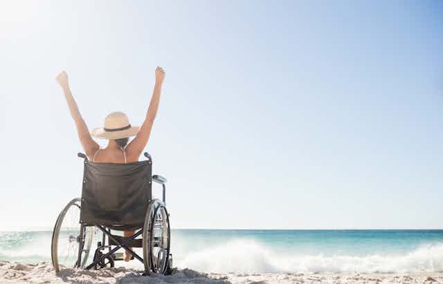 Woman in wheelchair raises arms in celebration on a sandy beach with crashing waves.