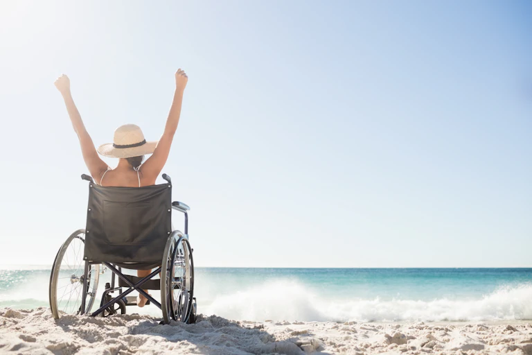 Woman in wheelchair raises arms in celebration on a sandy beach with crashing waves.