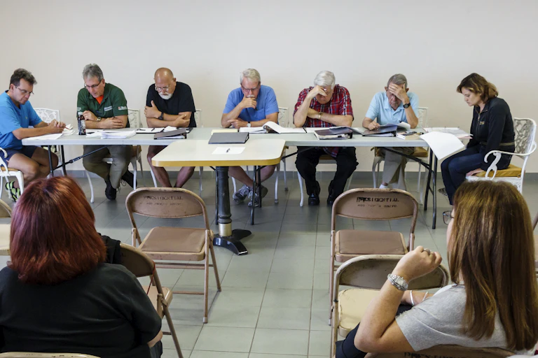 People attend a meeting, with several of them lined up at the back of the room