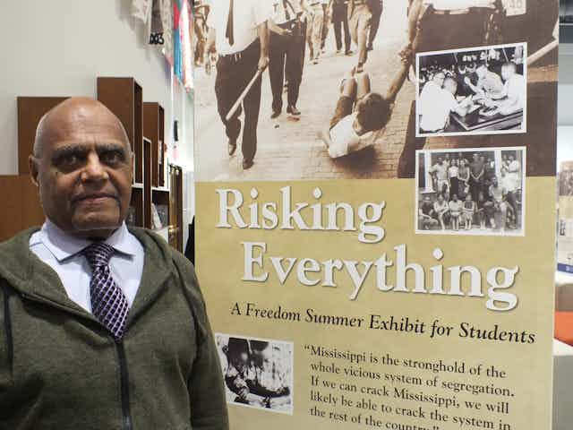 A Black man stands next to a poster which reads "Risking Everything".