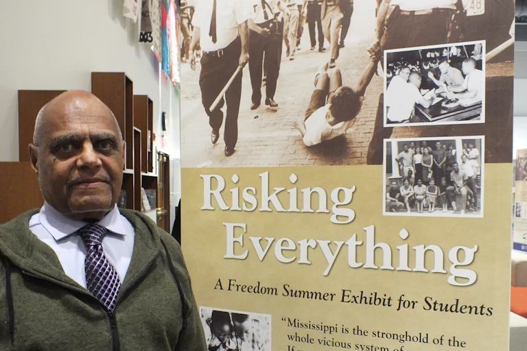 A Black man stands next to a poster which reads "Risking Everything".