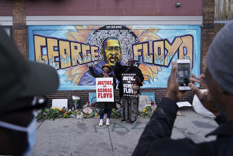 Two people stand holding signs that read 'Justice for George Floyd' in front of a mural depicting his face and name