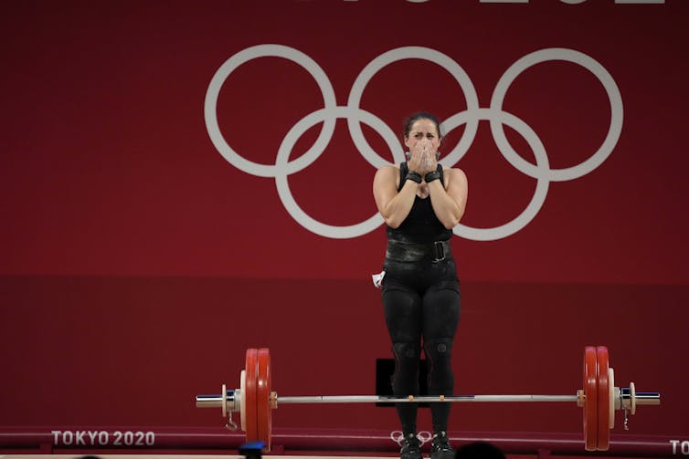 Woman standing behind weightlifting barbell covers the lower half of her face, looking overcome with emotion.