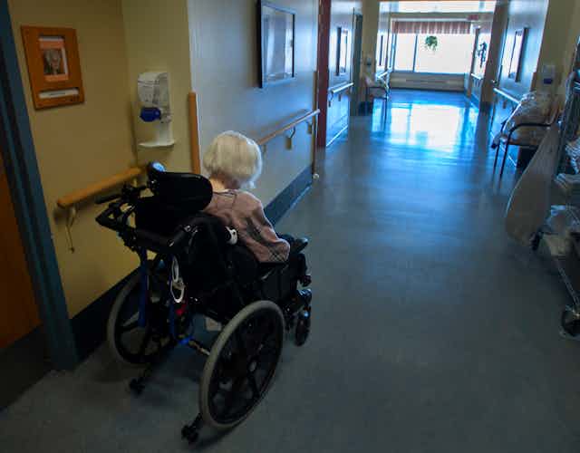 A grey-haired woman in a wheelchair seen from behind, in a long corridor