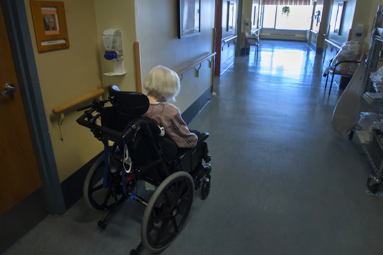 A grey-haired woman in a wheelchair seen from behind, in a long corridor