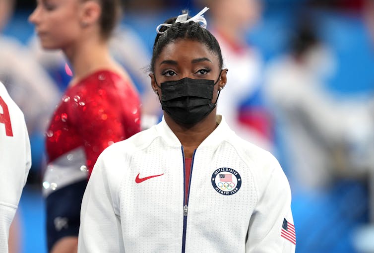 Simone Biles in a mask and white hoodie at the Olympics.