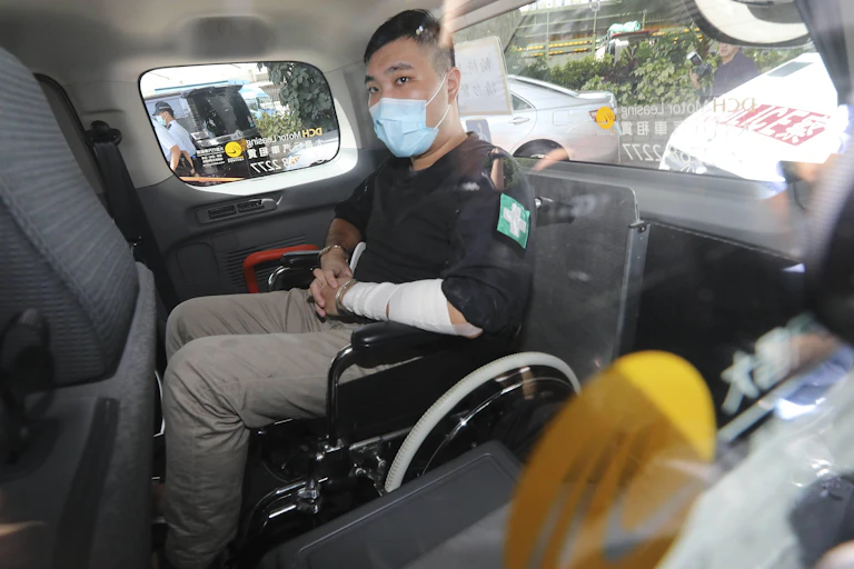 Tong Ying-kit, wearing a face mask, sitting in a wheelchair in the back of a van as he arrives to court.