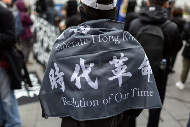 A protester draped in a grey banner with white text reading 'Liberate Hong Kong, revolution of our time.'