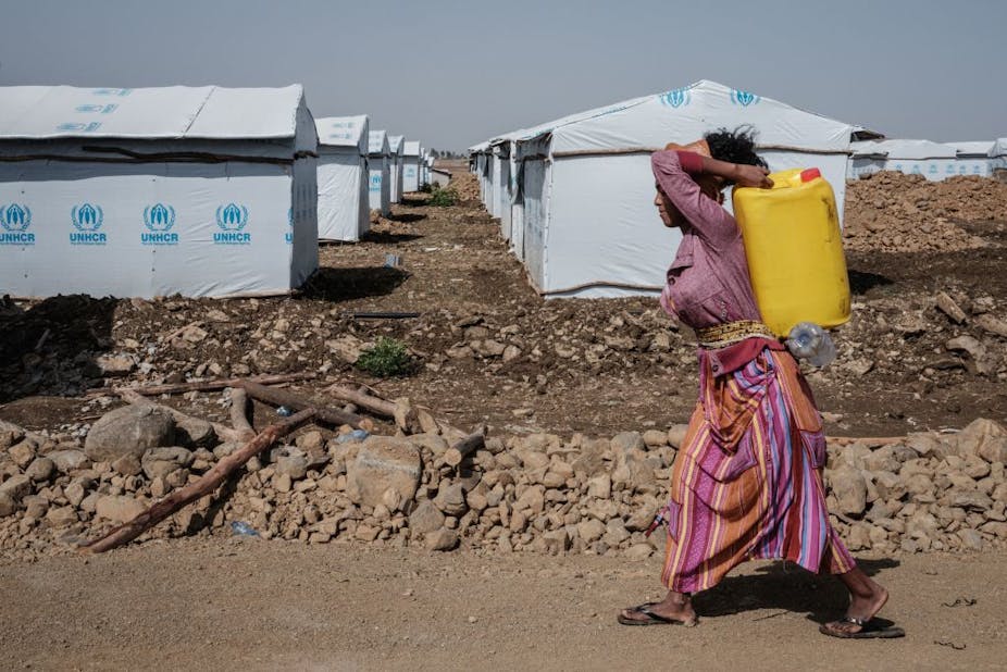 A woman carries a water container on her back as she walks past rows of tents