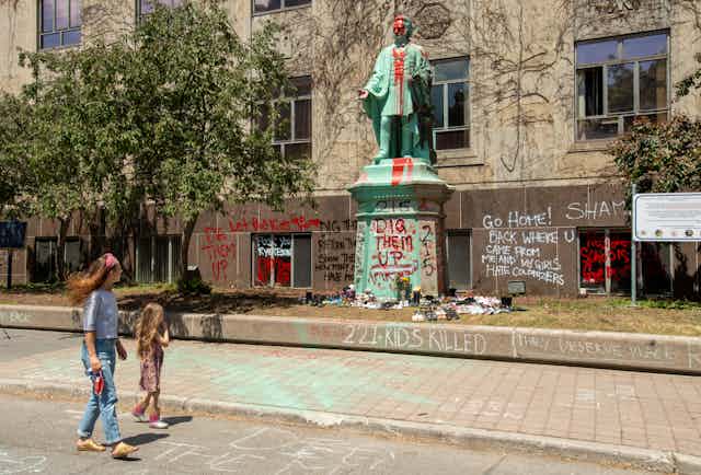A woman and child walk past shoes placed at the base of the vandalized Egerton Ryerson statue