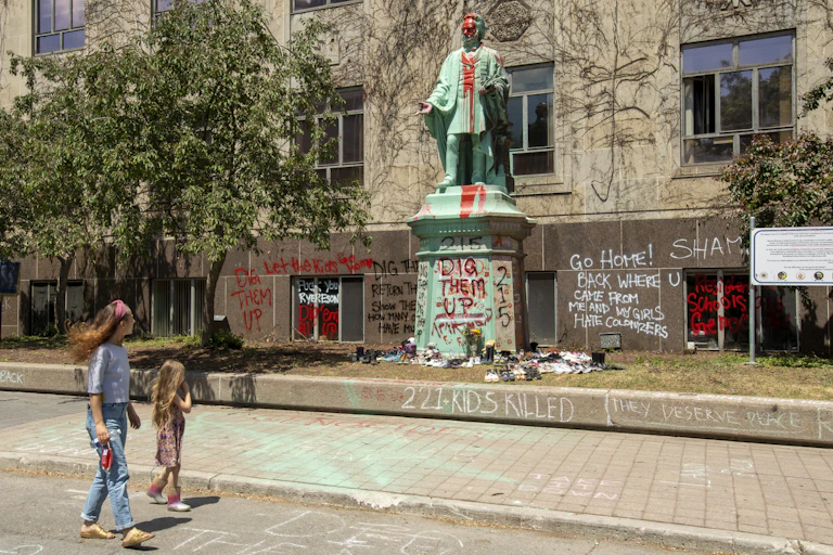 A woman and child walk past shoes placed at the base of the vandalized Egerton Ryerson statue