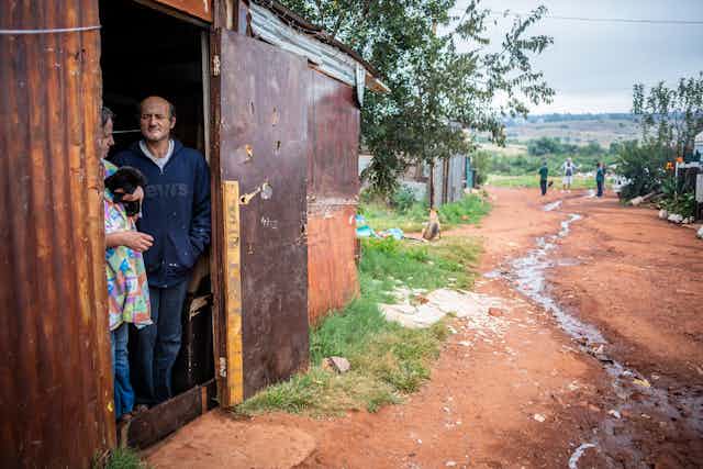 A woman and a man stand in the doorway of a shack, with waste water running down the unpaved road