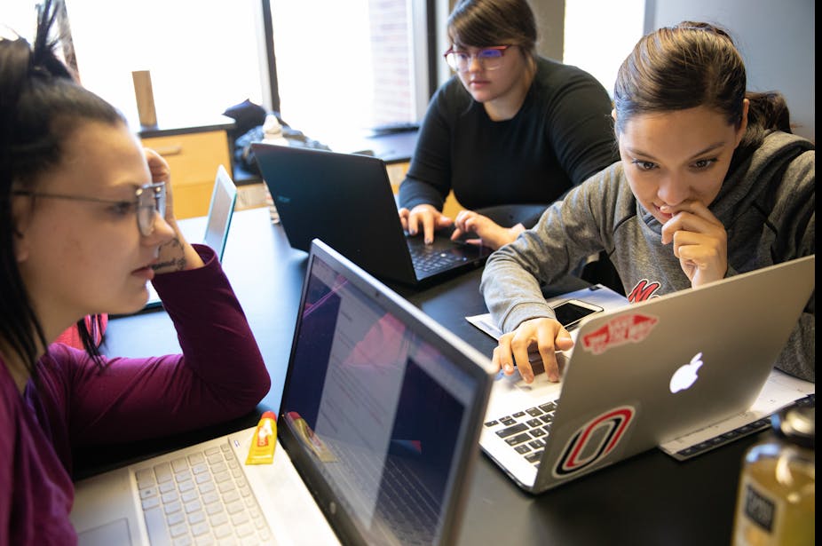 three young women sitting together at a table with their laptops open