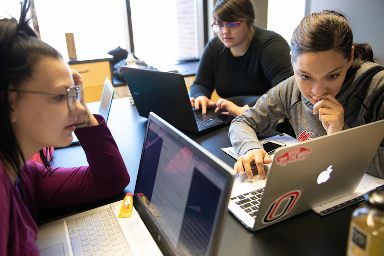 three young women sitting together at a table with their laptops open