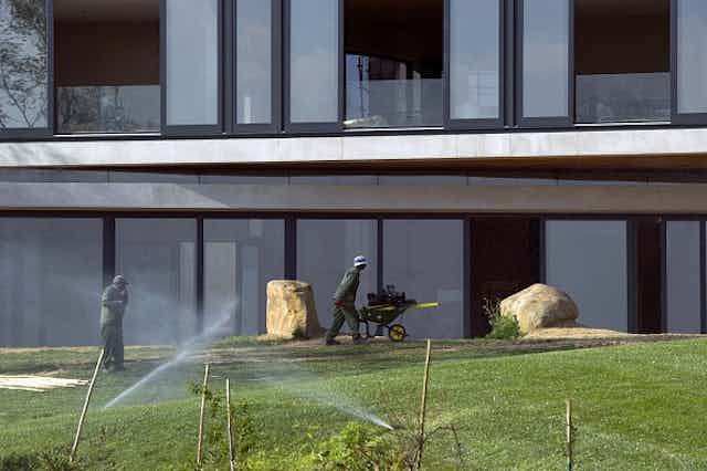 Man pushes a wheelbarrow past a building; sprinklers watering plants in the foreground