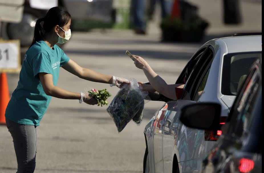 A masked woman hands bagged vegetables to a customer seated in a car.