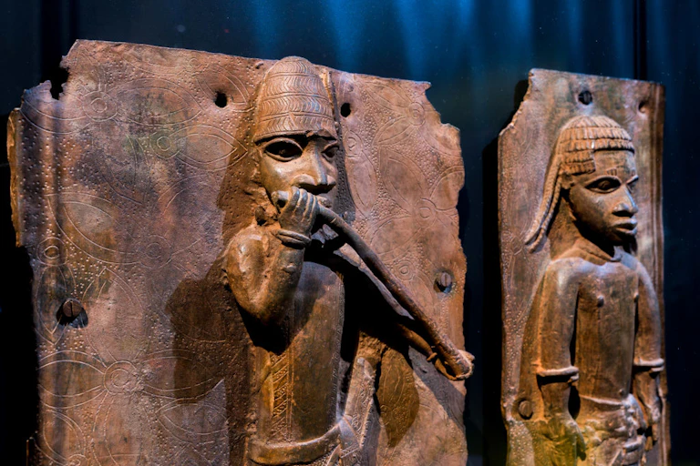 Metal sculptures against a dark curtain. They show two figures in ancient attire.
