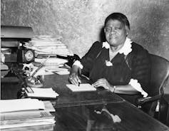 A woman sits at a desk surrounded by papers in an old photograph