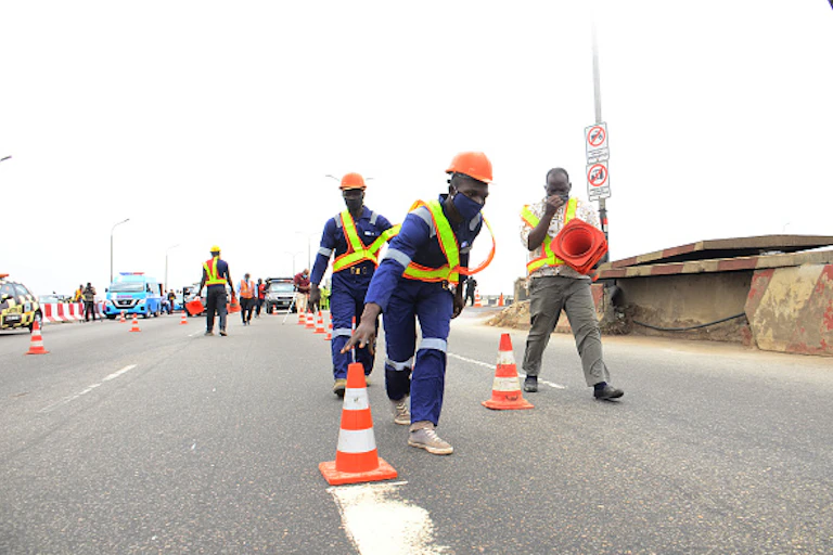 Construction workers on the road, one of them wearing mask adjusting the cone.