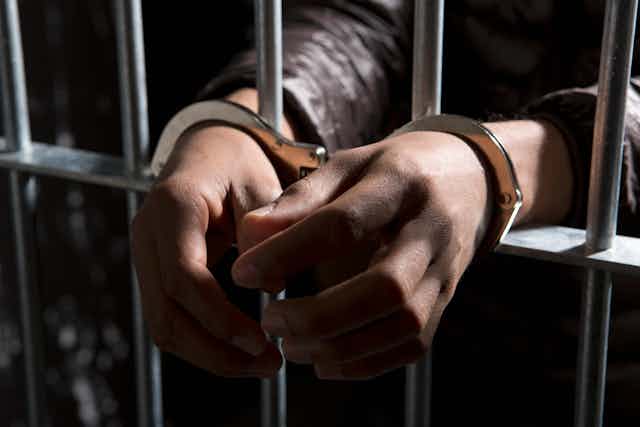 A person's hands pass through jail bars, with handcuffs around the wrists