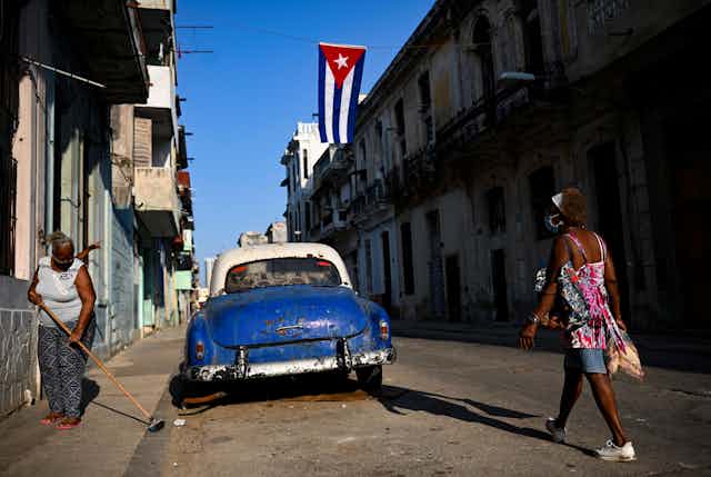 Street view with an old blue car, a woman sweeping the street and another woman walking down the street