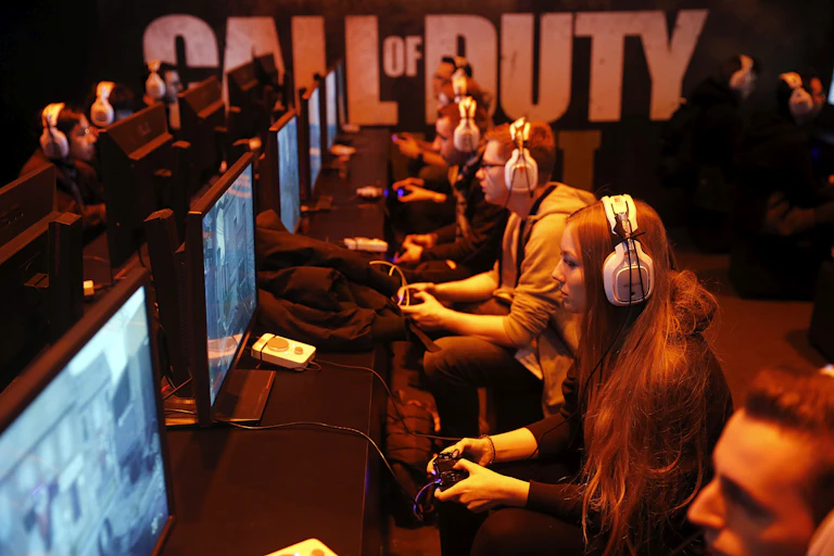 Several rows of young people wearing headphones and holding game control units stare at computer screens in a dark room