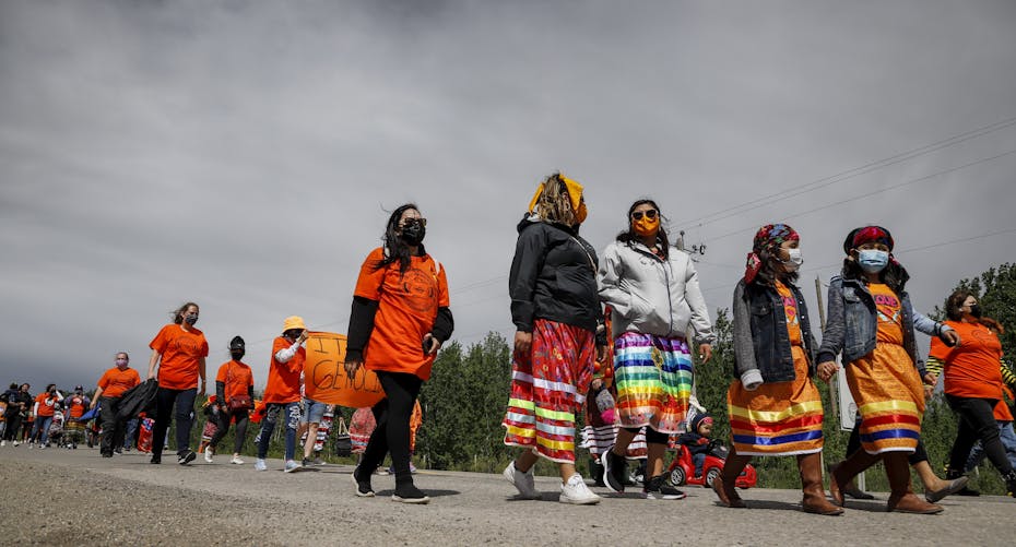 A group of people in orange shirts and ribbon skirts is seen walking down a road.