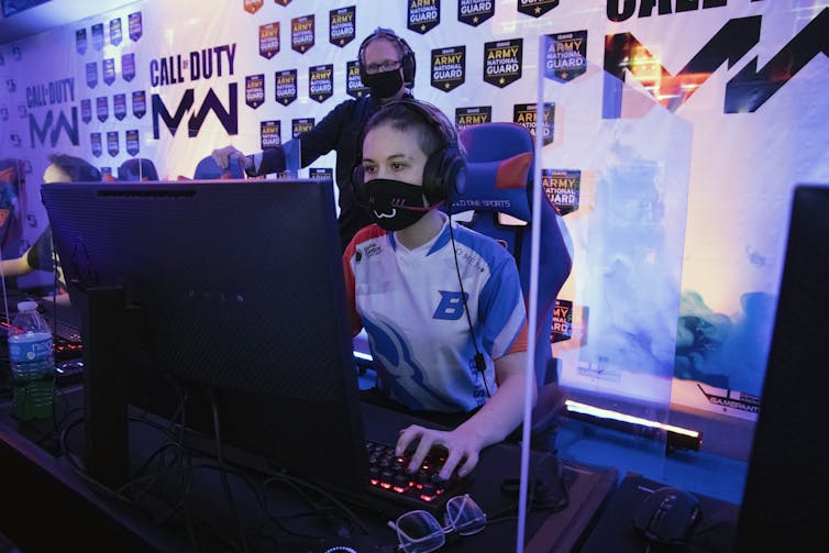 A young woman wearing a face mask stares intently at a large computer screen while a man wearing a face mask stands behind her looking over her shoulder