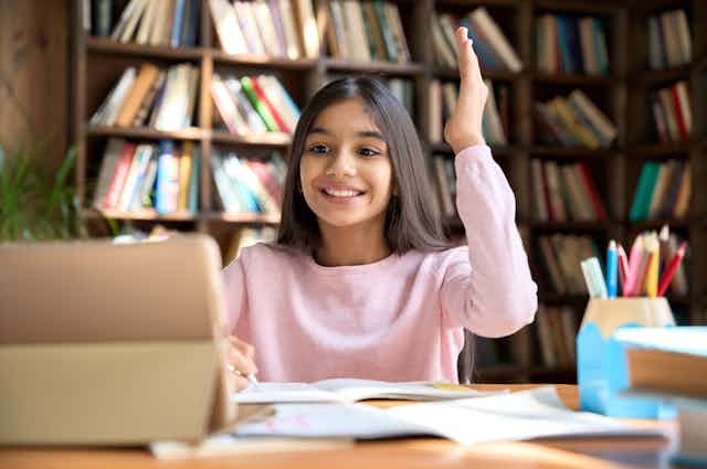 Happy girl student seated at desk in front of laptop raises hand 