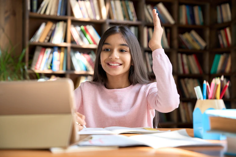 Happy girl student seated at desk in front of laptop raises hand