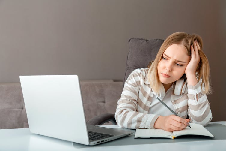 Worried looking teenage girl writing in notebook as she looks at laptop screen