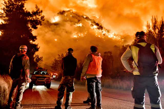 Firefighters look up at a hillside engulfed in fire.