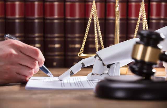 A human hand with a pen in hand at left signs a document where a robot hand points on a document with legal books in the background