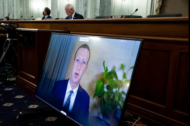 a large video screen with a man's face on it is propped against a large wooden bench behind which sit two men