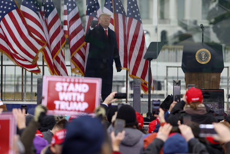 President Trump in front of flags and on a stage at a rally, with people crowded below him