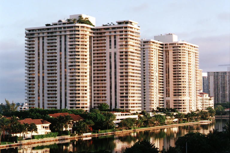 High-rise waterfront condominiums tower over tropical trees along the waterfront.