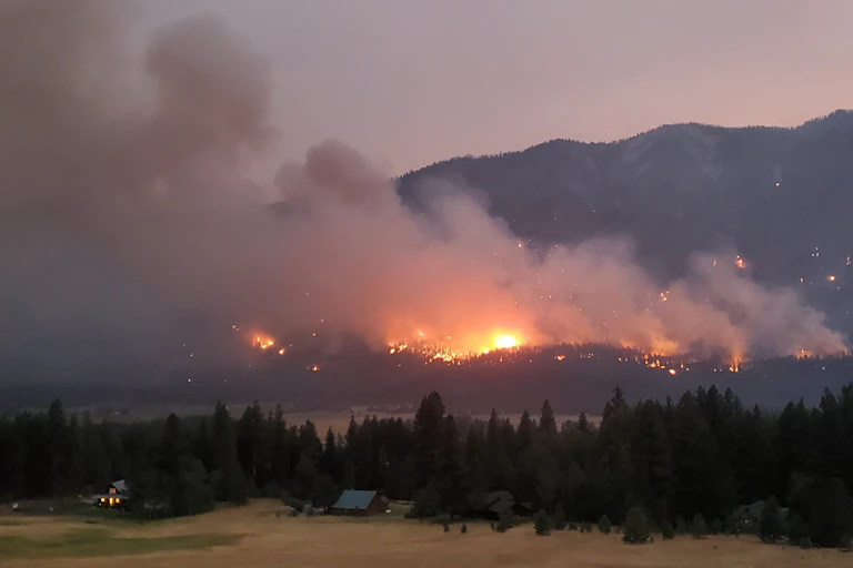 A fire burns in forested hills with homes in the foreground