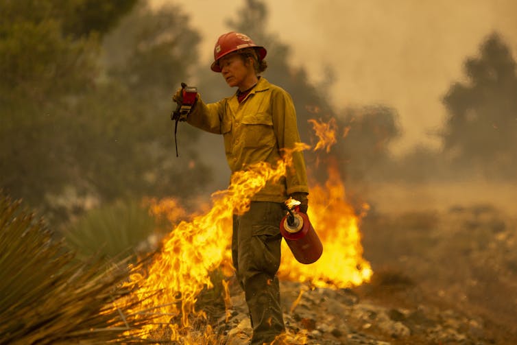 Una mujer bombero sostiene un walkie-talkie y una antorcha de goteo con una delgada línea de llamas detrás de ella.
