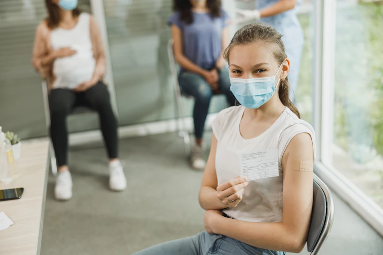 A young teenage girl displays her COVID vaccination card. Her upper arm has a band-aid.