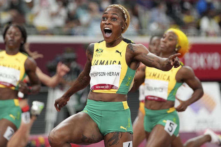 Thompson-Herah reacts with joy with her arms outstretched while her teammates cross the finish line in the background.